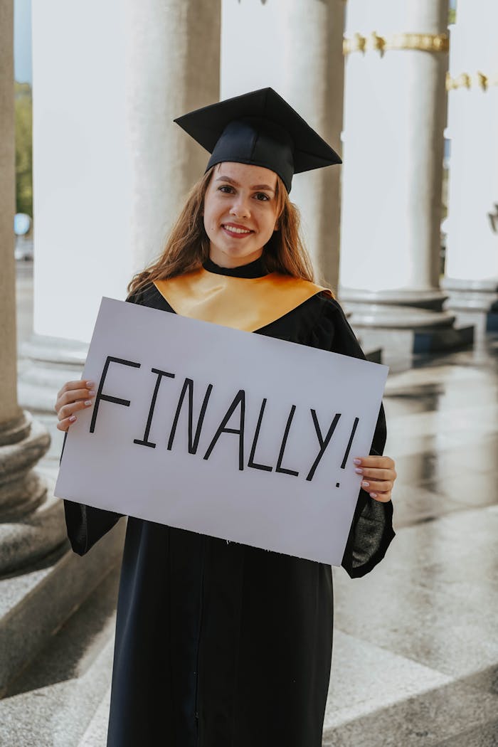 Young woman in graduation gown smiling and holding a u0022Finally!u0022 sign outdoors.
